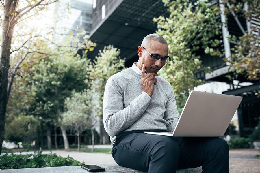Blog - Portrait of a Young Businessman Sitting Outside of an Office Building Surrounded by Green Foliage Using a Laptop