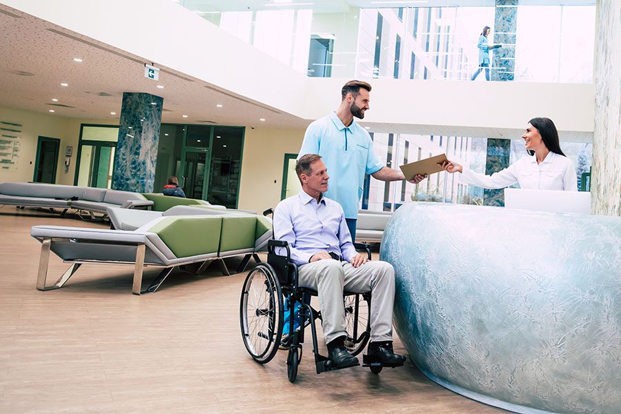 Specialized Business Insurance - View of Senior Man in a Wheelchair in the Hospital Being Attended by a Member of the Medical Staff in the Reception Area
